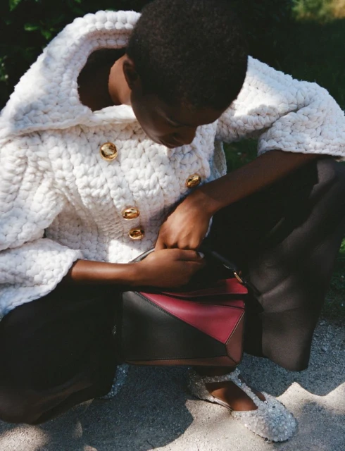 Woman in a white knit jacket with gold buttons, black pants and embellished shoes, holding a burgundy and black geometric handbag outdoors.