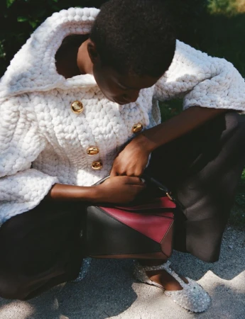 Woman in a white knit jacket with gold buttons, black pants and embellished shoes, holding a burgundy and black geometric handbag outdoors.