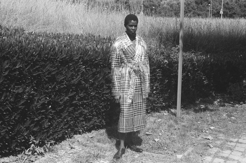 A person in a patterned coat stands on a lawn with a hedge in the background, creating a monochromatic outdoor portrait.