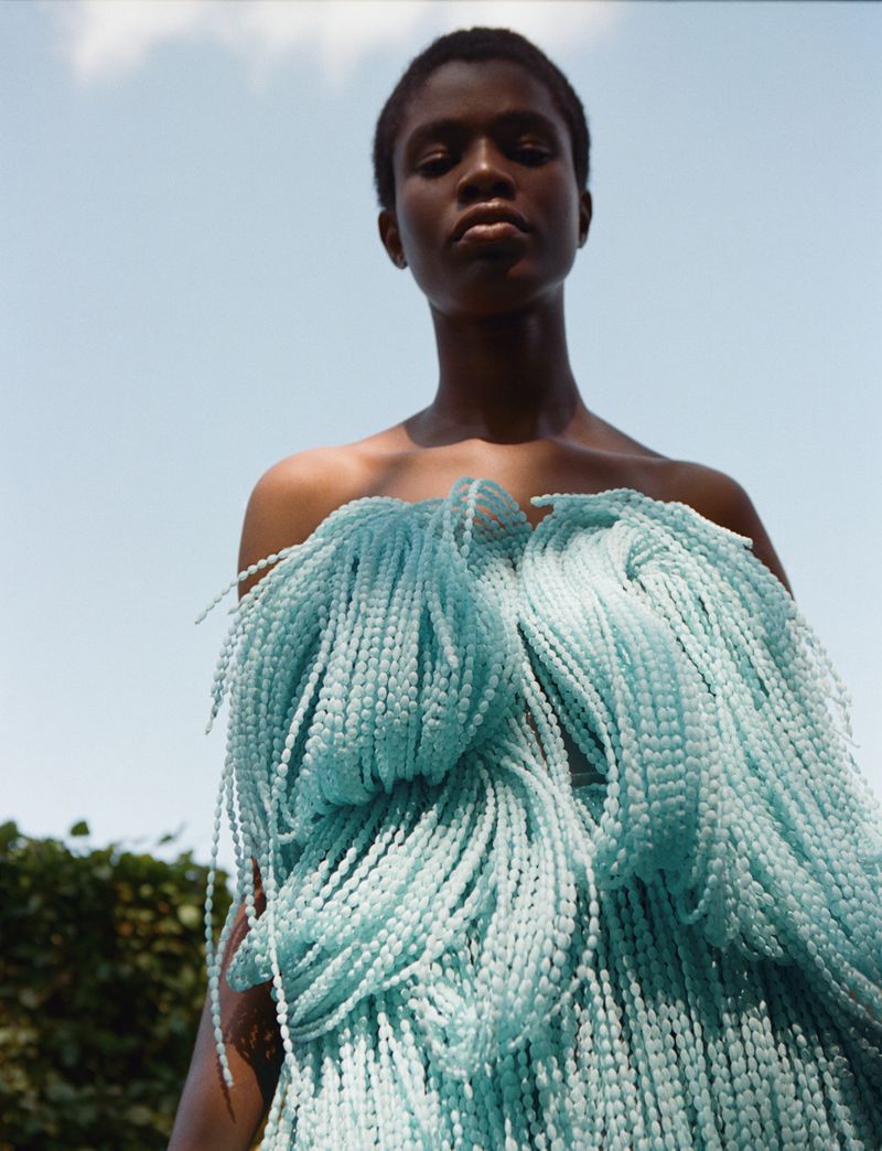 Adult female poses outdoors wearing an aqua-colored fringed garment with sky and foliage background in a medium shot at eye-level.