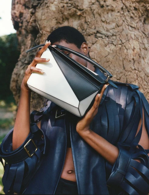 Woman showcasing a black and white geometric handbag in an outdoor fashion shot, wearing a dark, fringed leather jacket, tree backdrop.