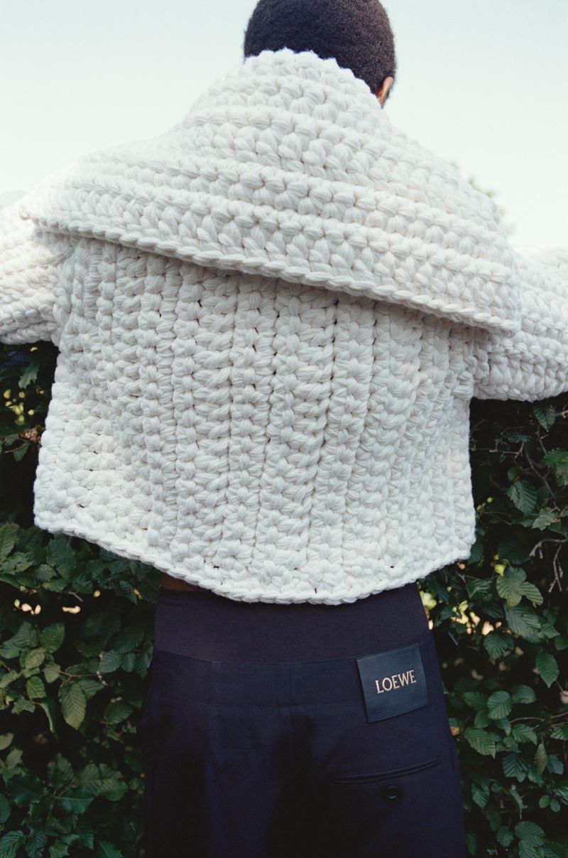 Back view of a person wearing a chunky, ivory-colored knit sweater with dark pants and a Loewe brand tag in a natural setting.