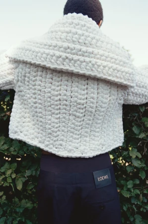 Back view of a person wearing a chunky, ivory-colored knit sweater with dark pants and a Loewe brand tag in a natural setting.