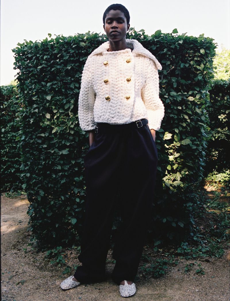 Fashionable African woman poses in a chunky knit jacket and wide-legged trousers, against a green hedge in an outdoor setting.