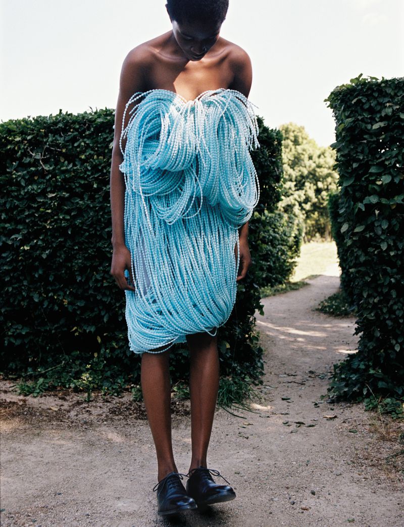 Black woman in a pale blue beaded dress standing in a manicured outdoor garden with green hedges and a gravel path. Fashion editorial.