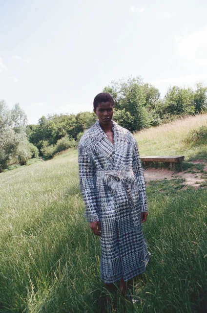 Dark-skinned adult stands outdoors in a field of tall green grass, wearing a patterned coat and a light-colored tie on a bright sunny day.