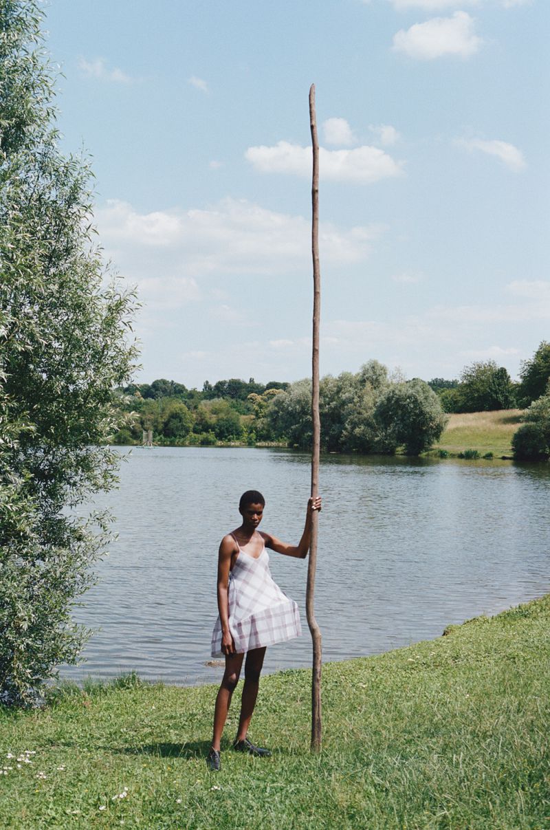 A woman in a dress is standing in a grassy field next to a lake, holding a tall wooden pole against a backdrop of trees.