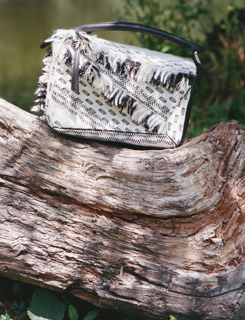 Fringed purse with geometric patterns resting on a fallen tree trunk in an outdoor setting with soft, natural light and green foliage.