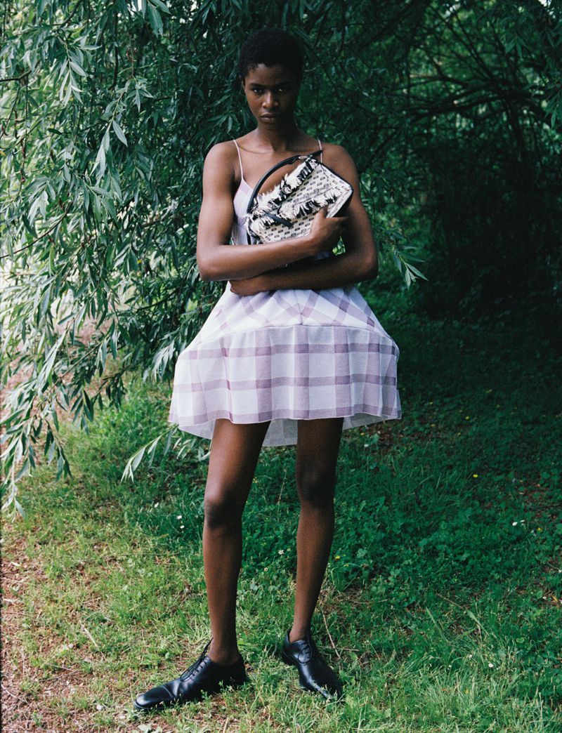 A woman in a dress stands in a grassy outdoor setting holding a purse, creating a natural fashion portrait.