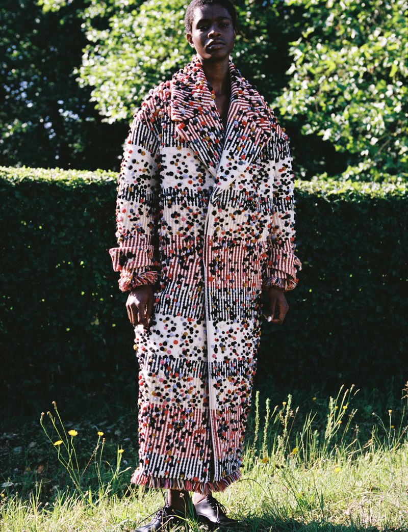 Model Onyinyechi Chijioke standing outdoors in front of a lush hedge wearing a long, multi-textured fringed statement coat.