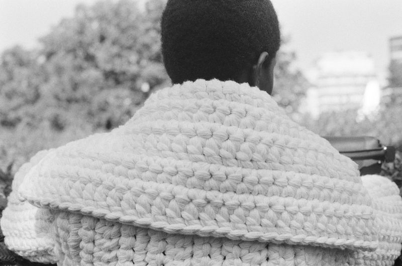 Person wearing a white crocheted poncho outdoors, back view, with trees and an urban building visible in the background, black and white.