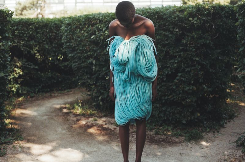 Adult stands wearing a dress of braided blue cords, photographed in a green outdoor setting with a meandering dirt path.