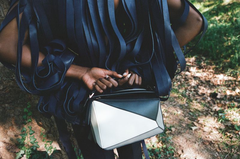 Stylish woman wearing a blue ribbon dress with a color-blocked geometric bag, standing outdoors amongst green foliage and a tree trunk.