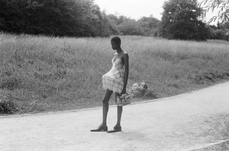 Black and white photo of an adult female with short hair walking on a path in a park, holding a decorative bag with trees in background.