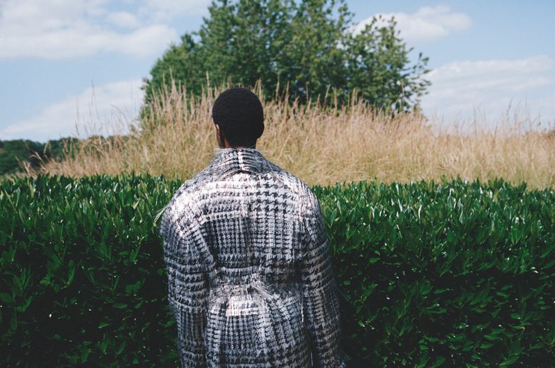 Person wearing a patterned overcoat stands with their back to the camera in front of a green hedge and grassy field on a sunny day.