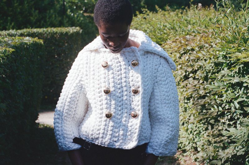 A woman stands outdoors wearing a white, double-breasted, textured jacket with large gold buttons, against a green hedge background.