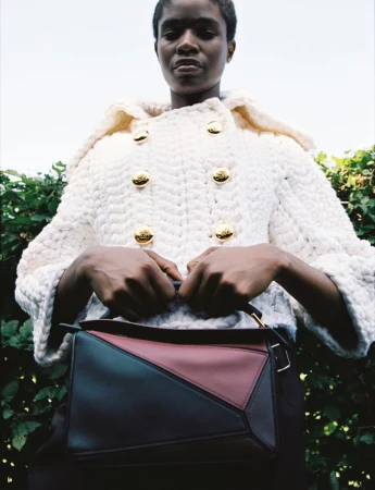 A dark-skinned woman models a cream-colored jacket with gold buttons and a tri-color Loewe handbag against a green hedge background.
