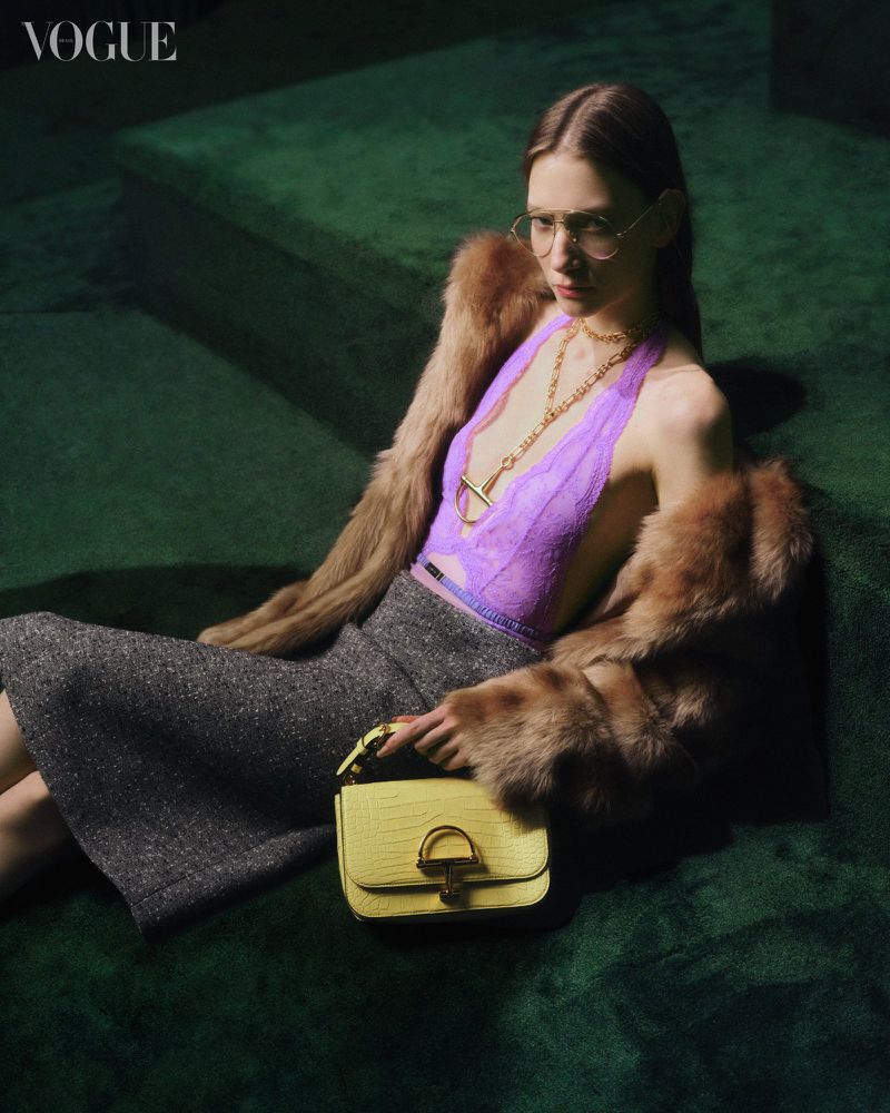 A woman poses on a green velvet surface with a fur coat and a yellow handbag for a fashion editorial, exuding elegance and style.
