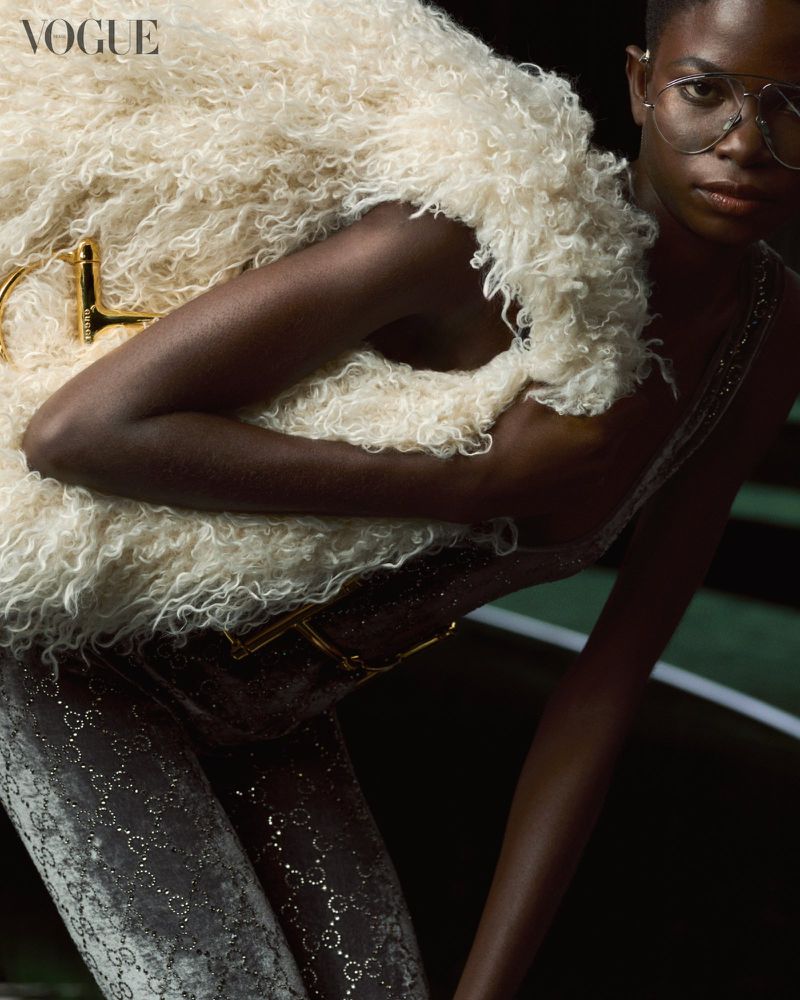 Fashion shot of an African woman wearing a fur vest and glasses with a patterned bodysuit in dark indoor setting, Vogue logo visible.