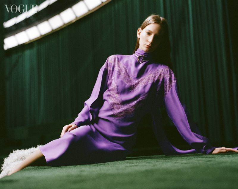 Fashion editorial shot of a model in a purple dress sitting against a dark green backdrop with soft, diffused lighting and Vogue logo.