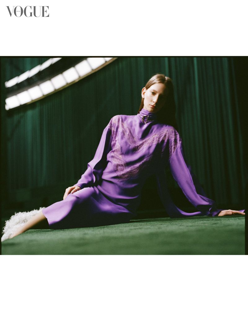 Fashion editorial shot featuring a woman in a long-sleeve purple dress, posed on a green surface with studio lighting and a Vogue logo.