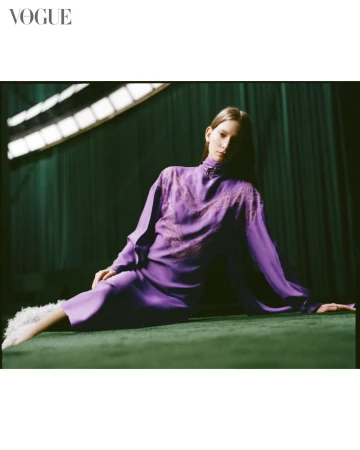 Fashion editorial shot featuring a woman in a long-sleeve purple dress, posed on a green surface with studio lighting and a Vogue logo.