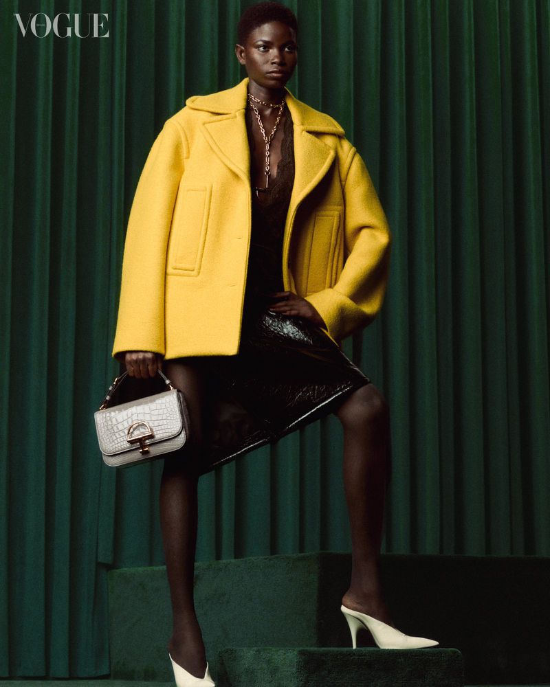 Fashion editorial shot of a Black woman in a yellow jacket, black dress, and white heels, posing with a structured handbag against a green backdrop.
