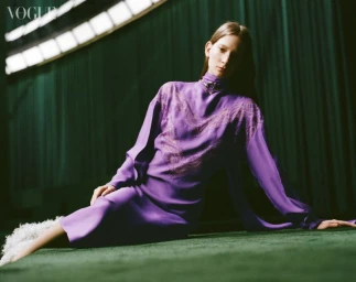 Model seated on a green carpet wearing a long-sleeved purple dress with metallic hardware against a dark green backdrop