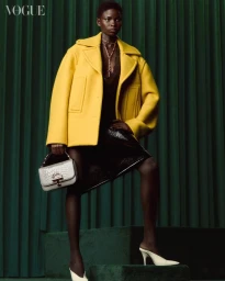 Model wearing a bright yellow jacket and holding a grey handbag against a pleated dark green backdrop.