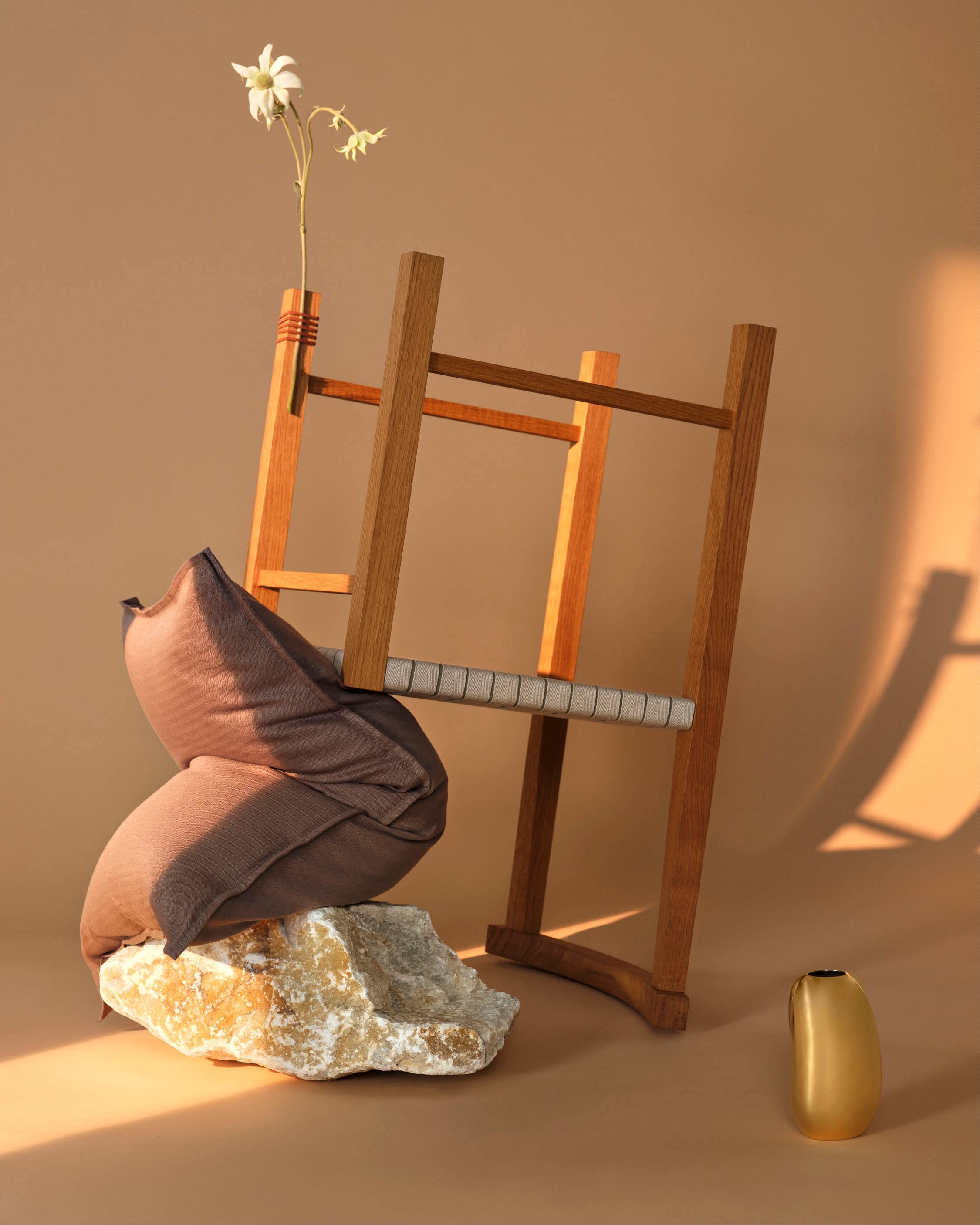 Still life shot of pillow on a rock, wooden chair, flowers in a vase, and small gold vase on a peach color seamless backdrop.