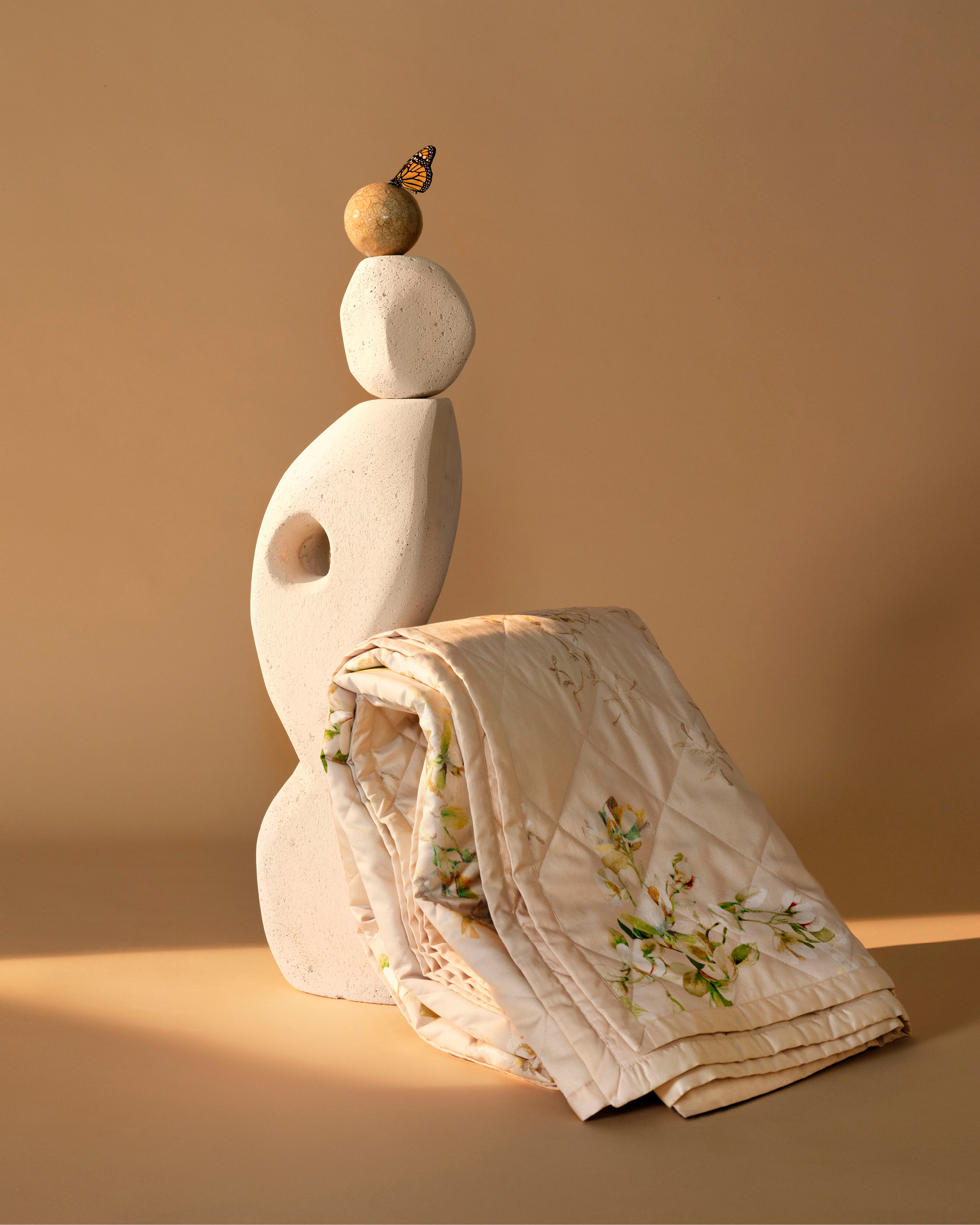 Still life with a butterfly perched on a stacked stone sculpture near a folded quilt with floral patterns in a neutral-toned setting.