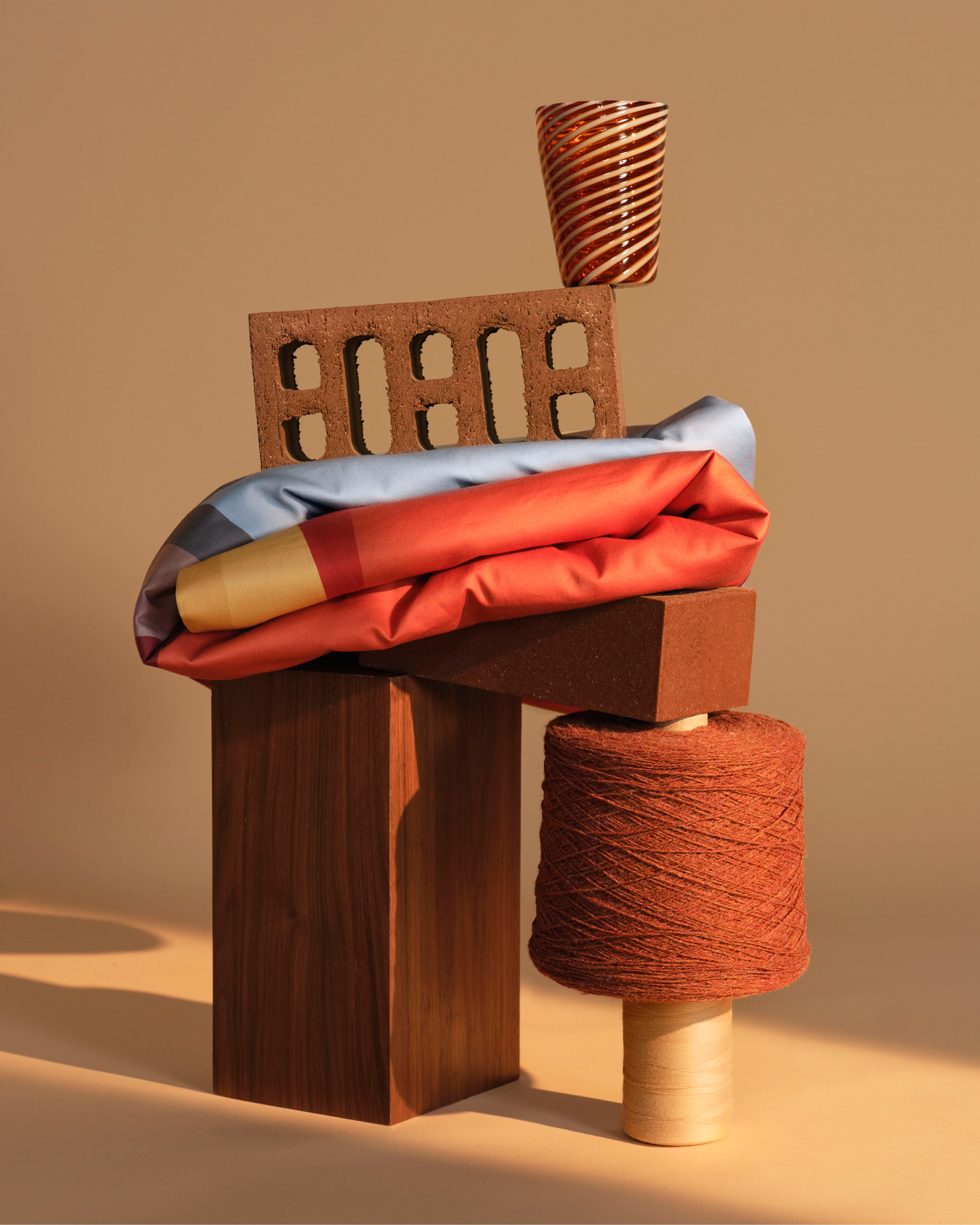 Abstract arrangement: brick, yarn spool, glass, draped textile, and wood blocks form a still life against a light brown background.