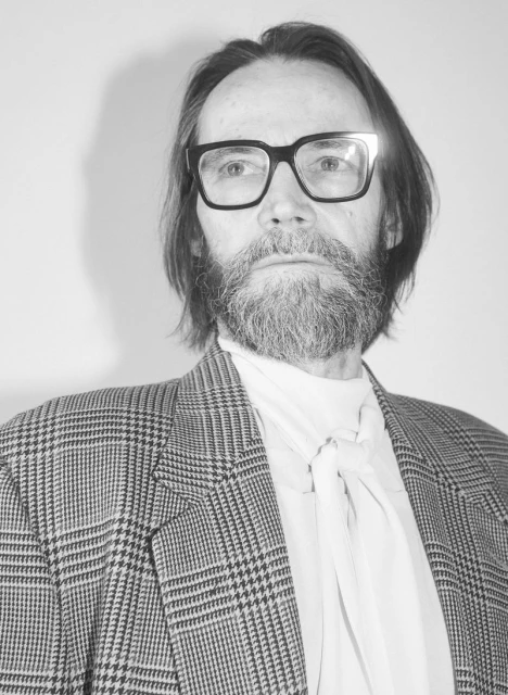 Black and white portrait of a bearded adult male wearing glasses and a patterned blazer, posing against a plain white background.
