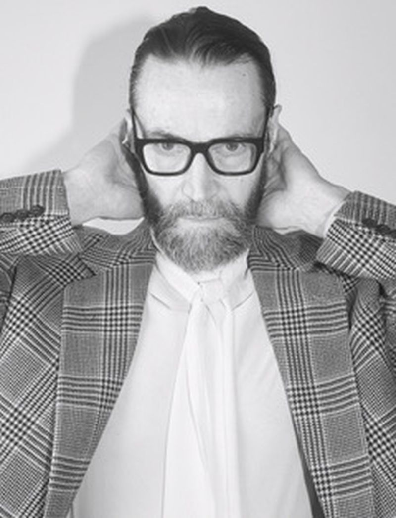 Black-and-white portrait of a bearded adult male wearing glasses and a patterned blazer, hands behind his neck against a plain background.