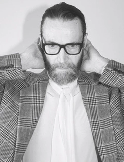 Black-and-white portrait of a bearded adult male wearing glasses and a patterned blazer, hands behind his neck against a plain background.