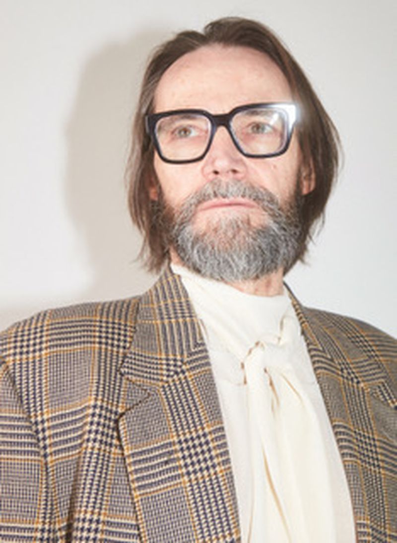 An adult man with glasses and a beard poses in a plaid blazer and cream-colored top against a neutral backdrop in a studio setting.