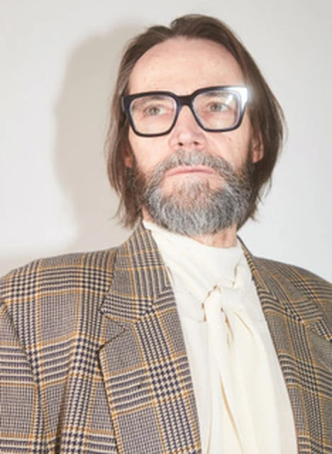 An adult man with glasses and a beard poses in a plaid blazer and cream-colored top against a neutral backdrop in a studio setting.