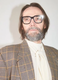 Karl Heinz Hirschberger wearing thick-frame glasses and a plaid blazer over a cream blouse against a white background.