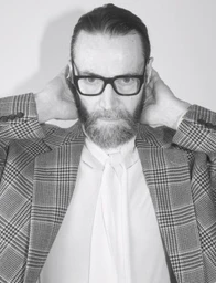 Black and white portrait of Karl Heinz Hirschberger in glasses and a plaid blazer with hands behind his head.
