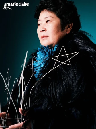 Portrait of an Asian woman with short black hair, wearing a dark coat and a blue top, posing with star-shaped props in a dark studio.