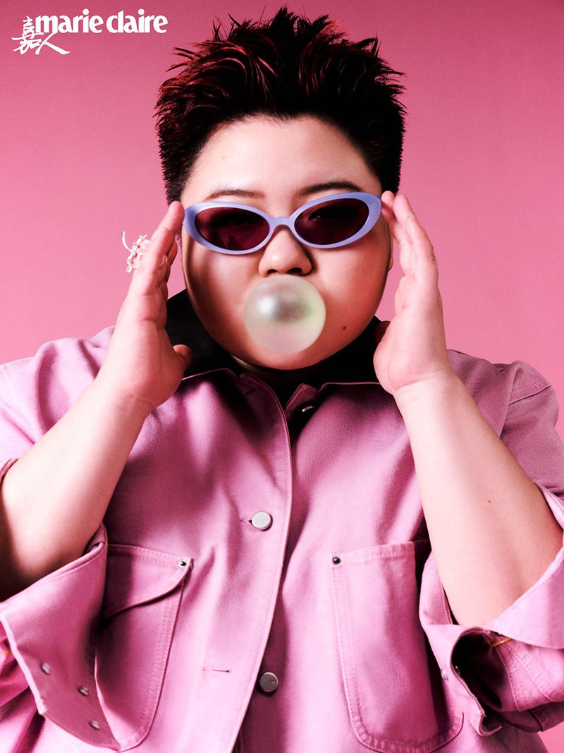Portrait of an adult female with short red hair wearing sunglasses and a pink jacket, blowing a bubble with bubblegum against a pink backdrop.