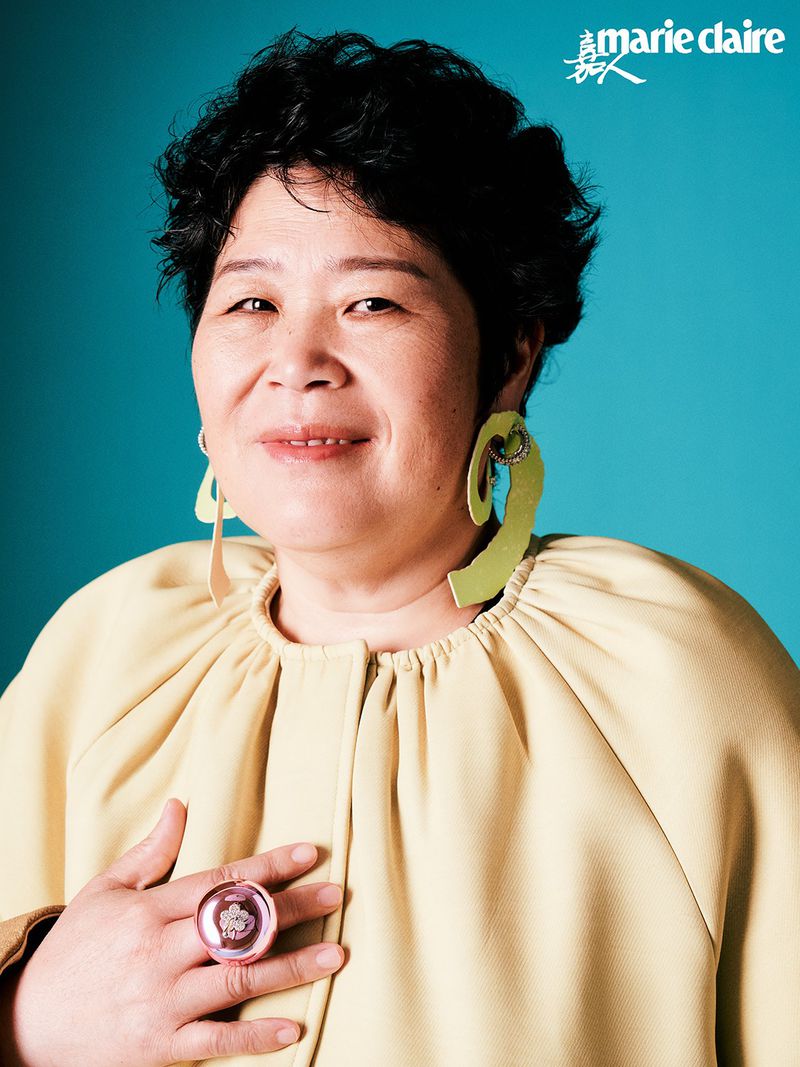 Portrait of an adult woman wearing a light yellow top and large green earrings against a turquoise background. She also has a big ring.
