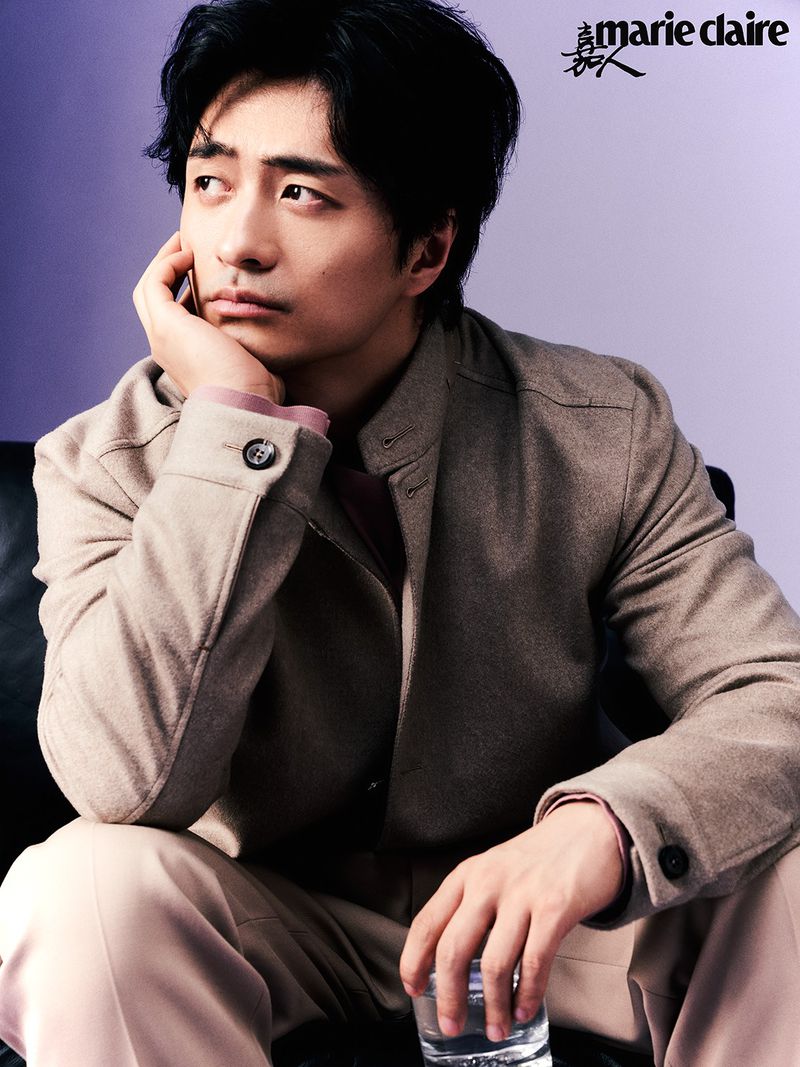Portrait of Asian man in jacket sitting with glass of water in hand, shot in photo studio with purple backdrop and soft diffused lighting.