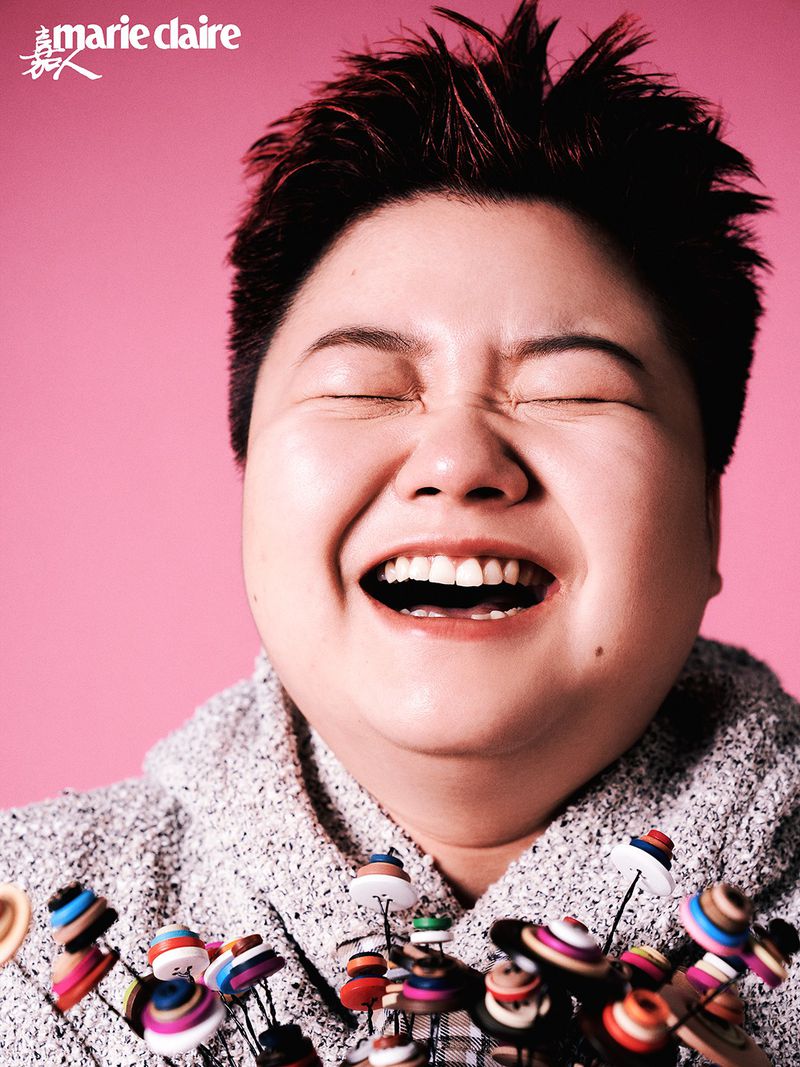 Close-up portrait of a smiling adult with short red hair, wearing a gray sweater and standing in front of a pink background. 