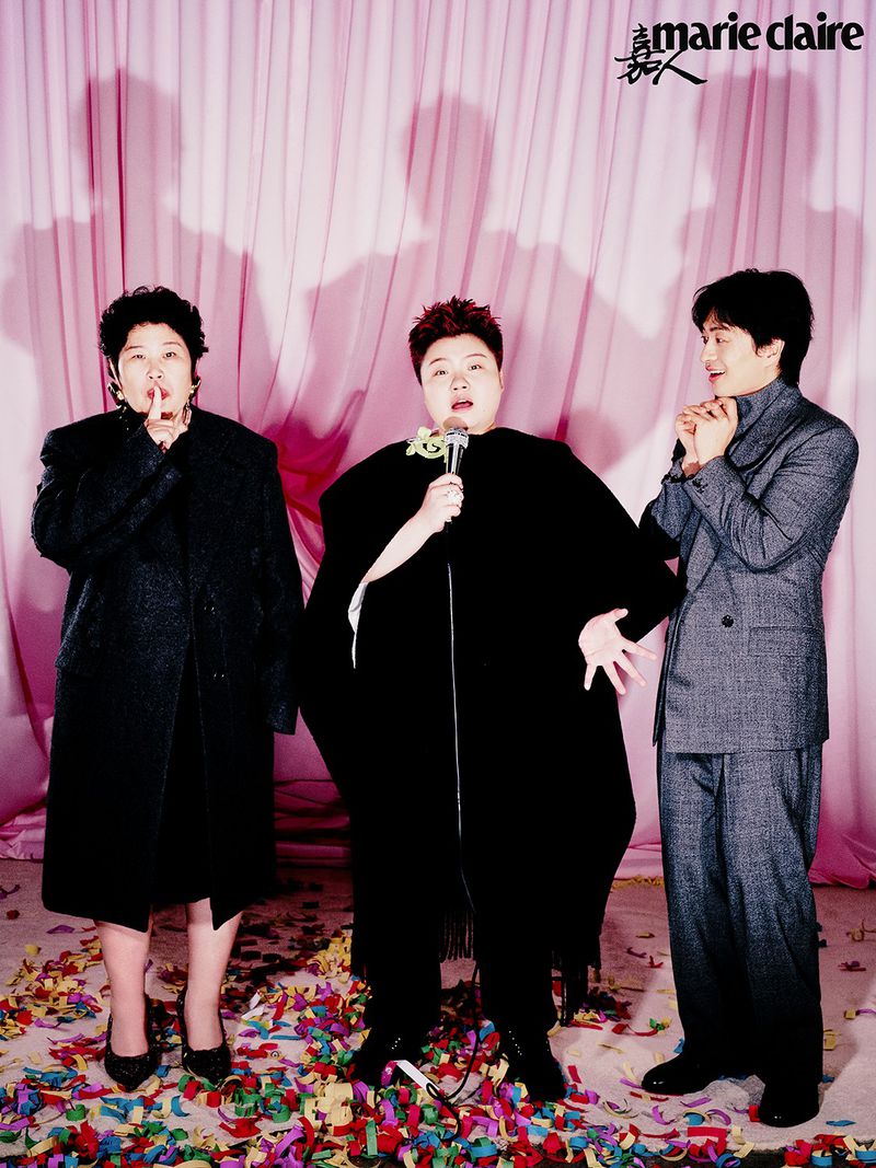 Three figures pose in front of a pink curtain with confetti: an adult female in a coat, a performer with a microphone, and a man in a suit.
