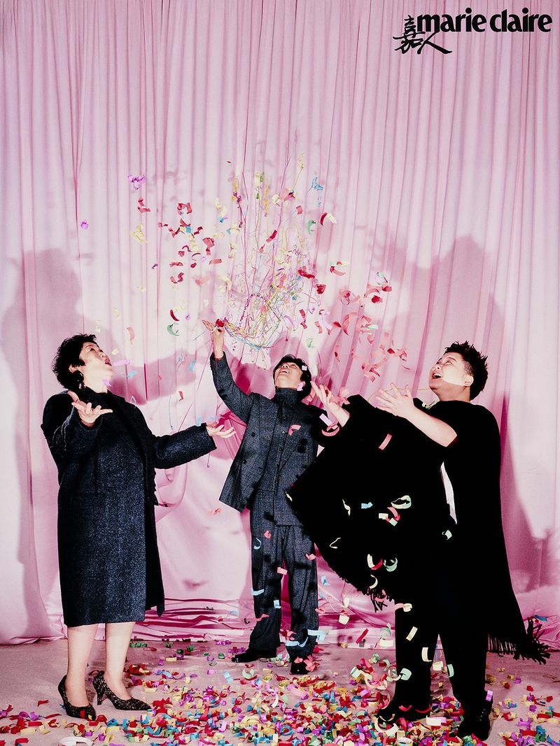 Three adults standing indoors with a pink curtain backdrop, smiling and throwing confetti, in a celebratory fashion editorial style image.