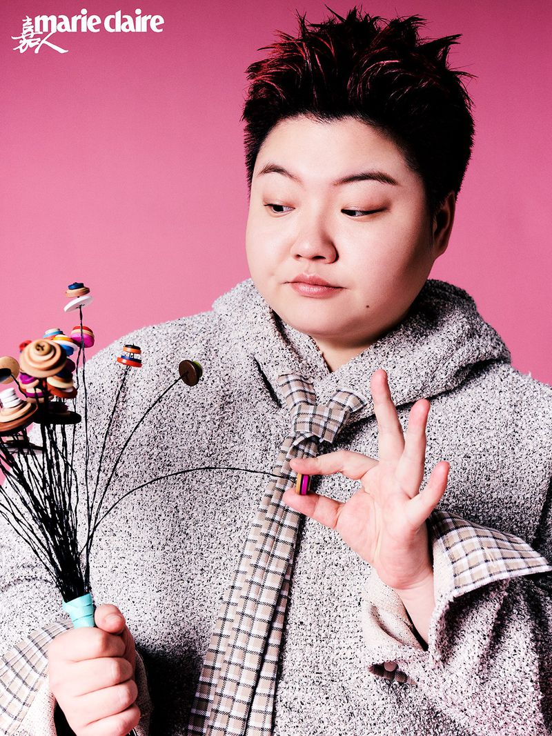 Portrait of a person holding a whimsical button bouquet, wearing a textured grey coat and patterned tie against a soft pink backdrop.