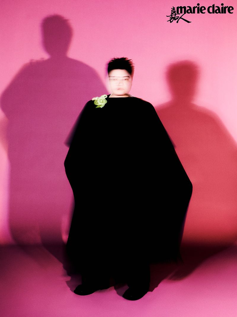A person in a black gown with a flower brooch stands in front of a pink background with blurred shadow figures, and the Marie Claire logo.