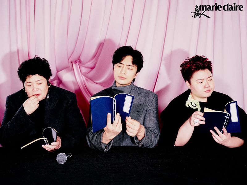 Three figures seated at a table against a pink backdrop, engrossed in reading blue-covered books in a studio setting.