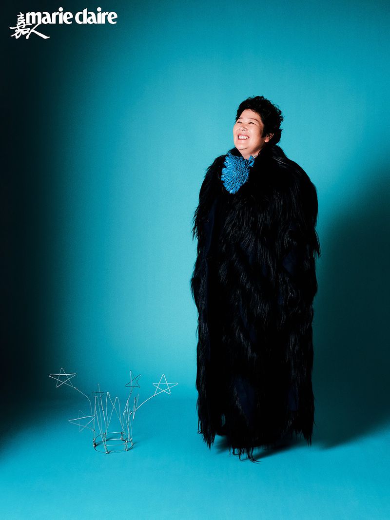 Smiling woman poses in a feathered black coat against a turquoise backdrop in a fashion editorial portrait with studio lighting.
